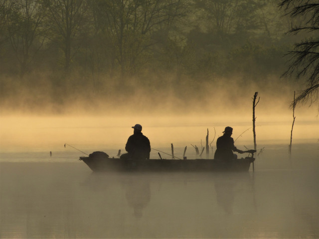 Photo of two men fishing from a small boat. They and the boat are in silhouette. The lake on which they float is misty and beginning to be lit by the morning sun, though they are still in the shadows.