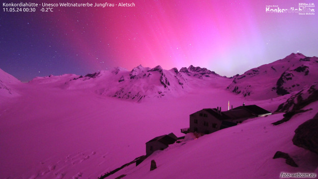 Nighttime view of the Konkordiahütte on the edge of the Aletsch glacier with the entire scene tinted pink by the light of the aurora.