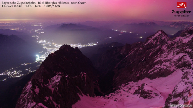 Nighttime view from the Zugspitze summit north over the dark Höllental and Garmisch-Partenkirchen with the entire scene tinted pink by the light of the aurora.