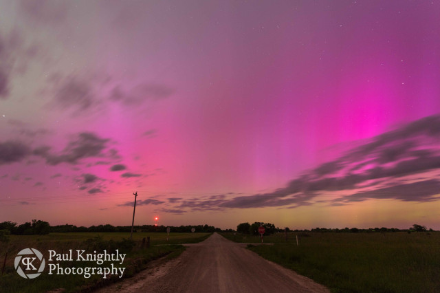 A road leads towards the vanishing point on the horizon beneath a purple and green aurora filled sky.