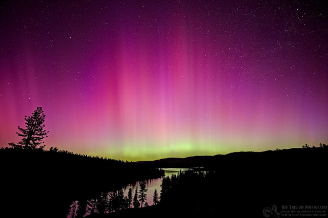 Color photo of an aurora. The landscape is forested mountains with a tongue of a lake going from the lower left to lower central frame. A distant body of water can be seen past the tongue. In the sky there are stars. On the horizon, just above the low mountain ridge line, is a band of yellow-green. Blue is above that. Then red. Then purple. There are numerous vertical streaks as well.