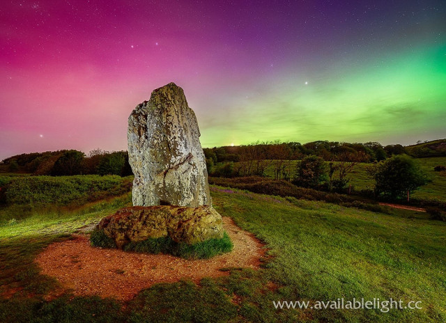 Vivid pinks, purples, blues, greens in the sky. A ancient standing stone in the foreground, illuminated by artificial light. A colourful and surreal image.