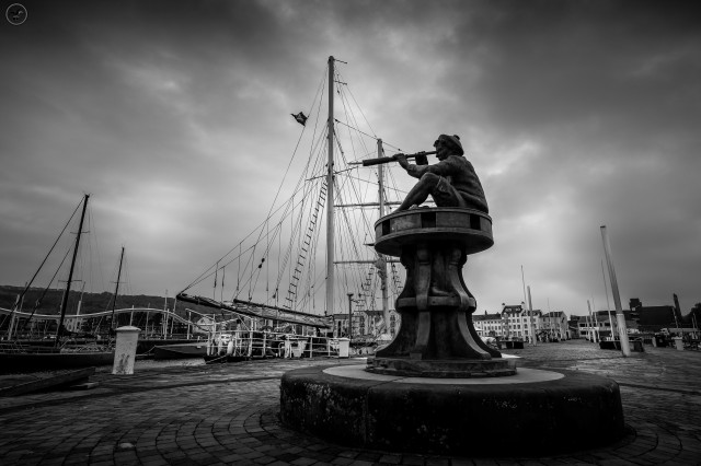 Monochrome shot of harbour statue of a boy with a telescope at end of stone jetty, with a large sailing ship in background and smaller boats around