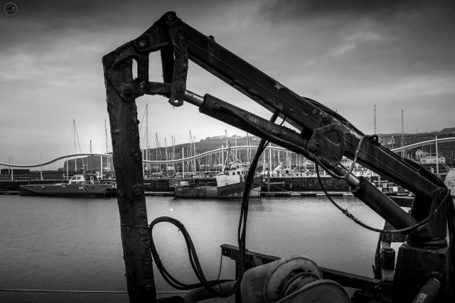 Monochrome shot through rusted crane jib with harbour in background, old sailing boat framed by crane jib