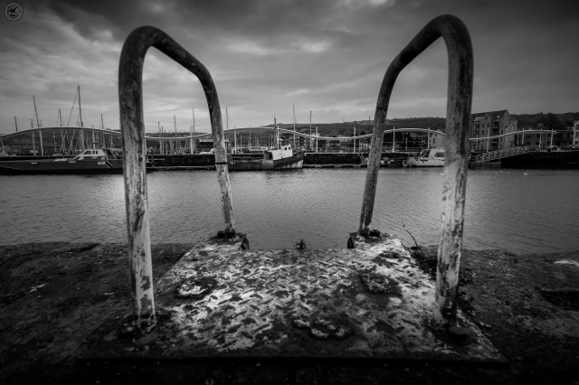 Monochrome shot through handles of mooring ladder with fishing boats in background