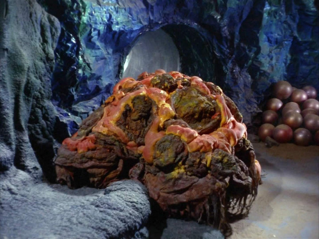 TOS scene. We're in a CAVE! In the background is a large group of circular balls that kind of look like a bunch of grapes but the size of basketballs. Front and center is a blob type creature that looks kind of rocky and also lava-ey. 