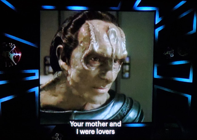 DS9 scene. Gul Dukat is seen on a computer monitor surrounded by 80's looking Tron like light zig-zags. He's a humanoid alien with a spiney boney facial structure and a spoon shaped forehead ridge. He looks slightly constipated. Closed caption reads, "Your mother and I were lovers."