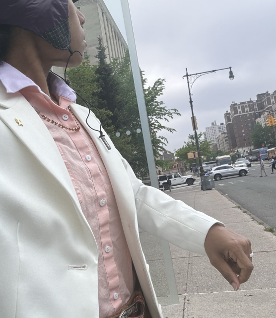 At the bus stop in a salmon linen shirt with lilac collar. White short cropped blazer, ultra light purple hunting cap. Gold pearl necklace and golden ant lapel pin. Traffic in NYC in the background. 