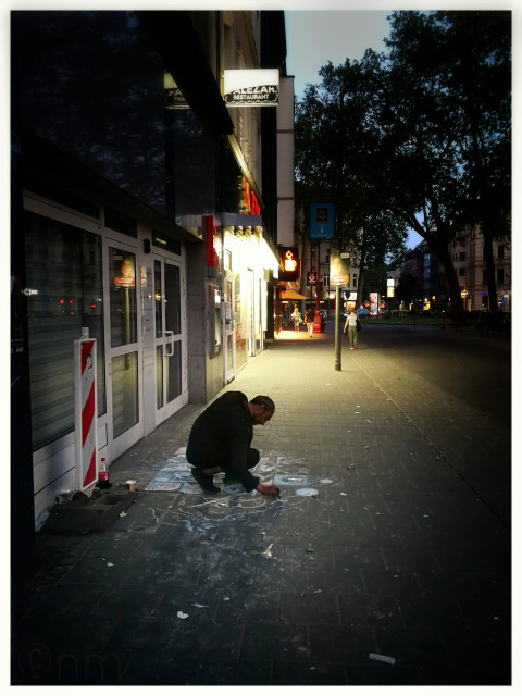 A homeless man dressed in black is kneeling on the sidewalk, drawing white chalk patterns on the concrete. The neon lights of an adjacent restaurant bathe the news ght scene in a yellowish light surrounded by a dark street, trees, and sky. 