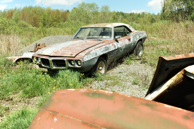 Sunbleached old muscle car (a Pontiac) lies in a field – gravel and grasses around it – hints of another car in the foreground – blue sky – clouds – peaceful feeling