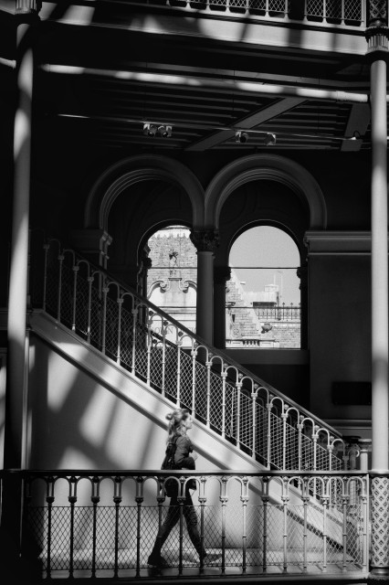 A black and white image of a person walking through a patch of light beside a staircase inside the National Museum in Edinburgh, with ornate railings and arches, and a glimpse of a building façade seen through a window in the background.