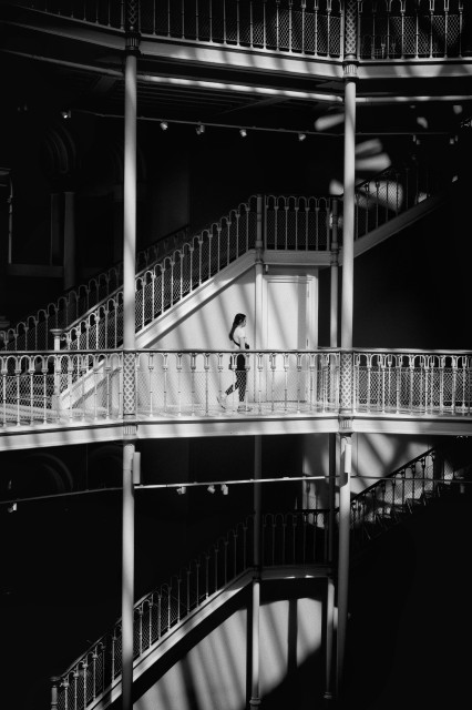 A young woman walks into a bright patch of sunlight on one of the upper floors of the National Museum in Edinburgh, where the ornate architecture is highlighted among dark, shadowed areas.