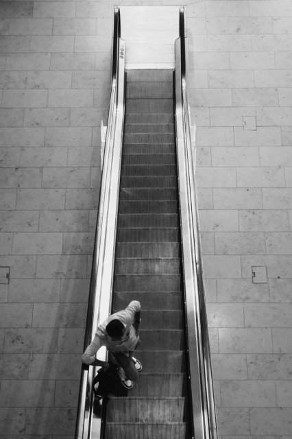 Black and white image of a person ascending an escalator, viewed from a high angle.