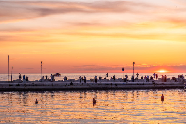 Ein Pier mit zahlreichen Menschen, die auf das offene Meer hinaussehen. Die Sonne verschwindet gerade am Horizont. Alles ist gelb-orange eingefärbt.