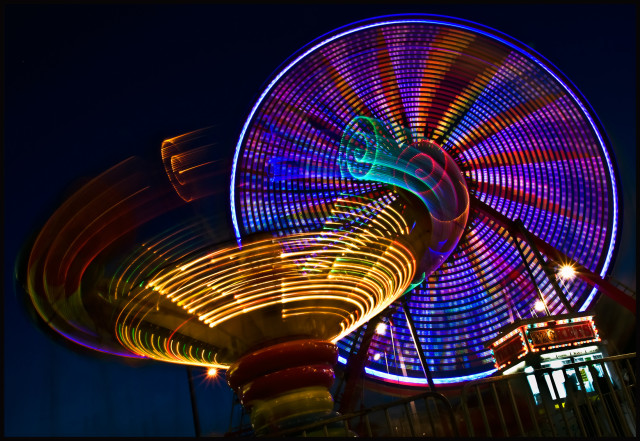 Two spinning carnival rides, photographed at night... brightly lit with colorful LEDs. 

The ride in the foreground is spinning like a cone-shaped top, lit in gold, teal, and red.

The ride in the rear is a giant ferris wheel with blue and red lights on the spokes.