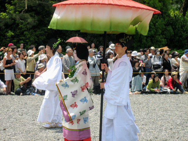 A female attendant, dressed in Heian period costume, is shielded from the fierce May sun by a giant parasol.