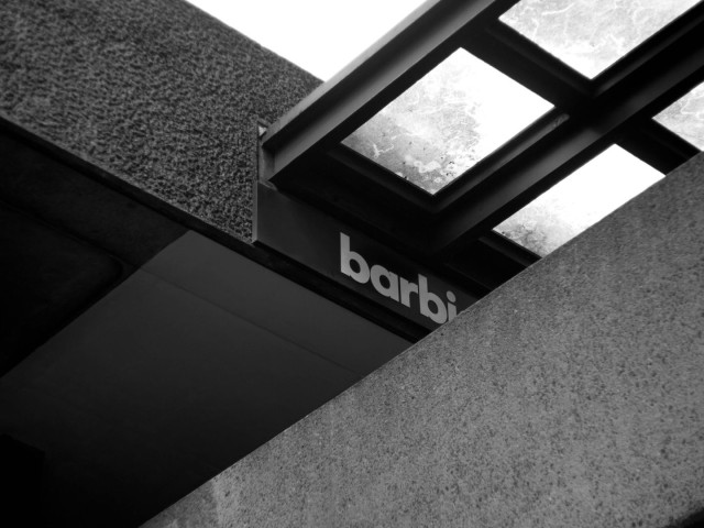 Monochrome photo of a section of a concrete walkway taken from below. The wall at the edge of the walkway takes up the bottom right corner, the building into which the walkway goes is at the left. Above the entrance is the beginning of a sign which says 'Barbi', the rest is obscured by the walkway wall. Above the sign is a rectangular porch or roof with glass panels and thick frames. There doesn't seem to be a single curved line in the picture.