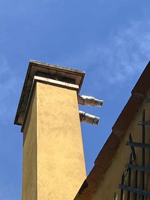 Colour photo looking up at a tall rectangular chimney, made of ochre coloured plaster, with two short silver slightly bulging ruched pipes sticking horizontally out of it which look very much like the arms of the robot from the 1960s TV series Lost In Space. The background is blue sky with a faint trail of white cloud. 