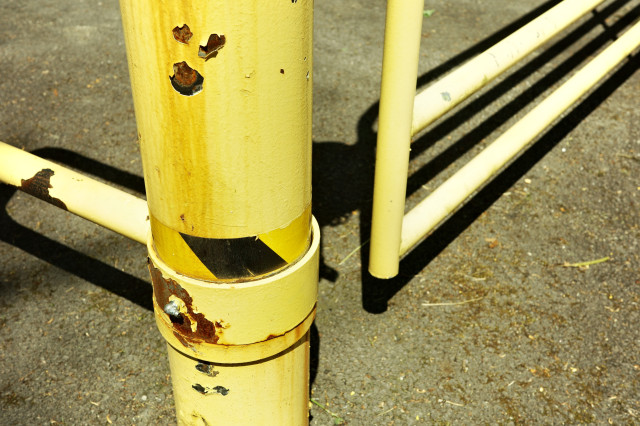 A stained yellow pole with black and yellow caution tape wrapped around its middle – peeling paint reveals rusted metal – yellow bars of a gate casting hard shadows on the concrete behind it