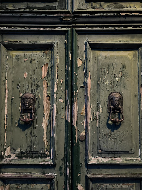 Colour photograph, taken in the gloaming half-light, of a pair of panelled wooden doors. They are painted a soft moss green colour, but the paint is cracked and chipped and peeling to an astonishing extent, revealing pale wood underneath. There is a symmetrical pair of door knockers in the shape of faces with what look like Egyptian headdresses, made of metal but it is impossible to see what type of metal under the tarnish and grime. To me the doors look welcoming and friendly, despite, or indeed because of, their shabby state, and I'd love to go and hang out in the presumably equally neglected Miss Havisham of a house that they open into, but I suspect that it might not be so appealing to someone with less finely-honed Goth-y sensibilities. 