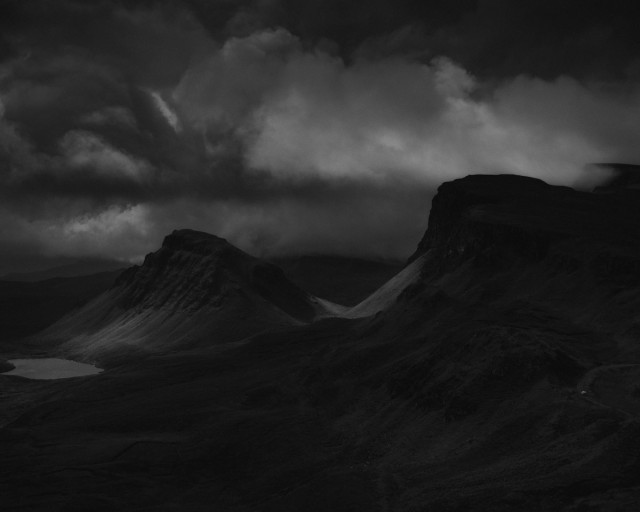 This image is a black and white photograph of dark mountain landscape from Scotland with dramatic stacked storm clouds towering over the primeval looking peaks and ridges sitting darkly below and lit in patches by broken sun. A small loch catches light on the far left. 