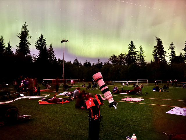 Sony A6500 with Sony 100-400mm lens on a ZWO AM 5 tripod mount in front of a yellow-green aurora. Taken at a star party with people in the background laying on the sports field and viewing the aurora.