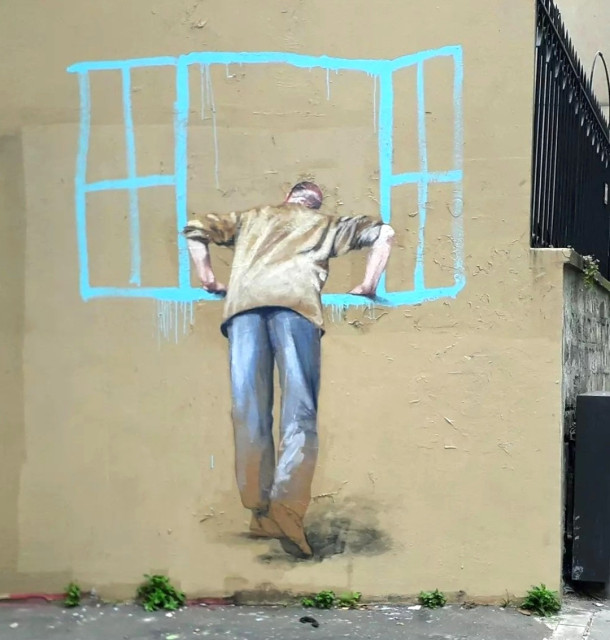 Streetartwall. A small mural with a man at a window has been sprayed/painted on a yellow exterior wall. The older man in jeans and a beige jacket is finely drawn and is reminiscent of a watercolor painting. The window, on the other hand, is indicated with a few light turquoise strokes and is reminiscent of a child's drawing. An imaginary window through which the older man is looking.