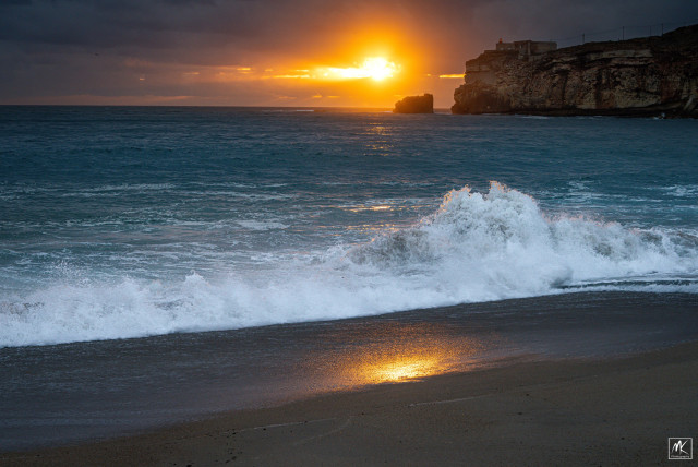 Color photo of a wave breaking on a beach, lit by the setting sun peeking through a hole in the clouds near the horizon. In the background to the right is a sheer cliff with a structure on top. 