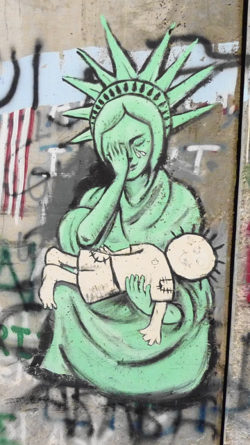 Palestinian graffito of the Statue of Liberty crying and covering her face with one hand, and cradling Handala with the other, reminiscent of Michelangelo's Madonna della Pietà.