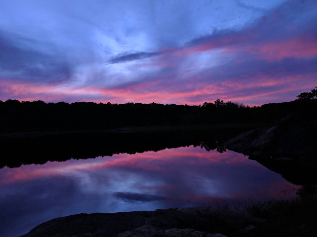 We are standing beside a lake. The rocky near shore and wooded far shore are each silhouetted in the deepening twilight. Against a pale blue sky a swirl of clouds is tinted dark pink and navy blue with touches of deep purple, mirrored by the lake below.
