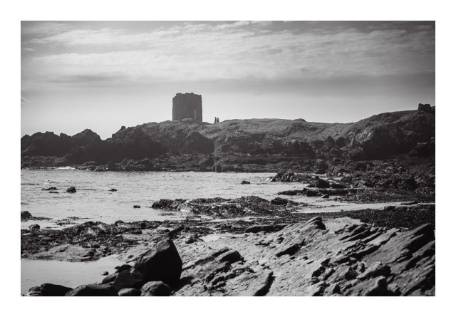 Black and white image of a rocky coastline with tidal pools, featuring a distant ruined tower and two people standing near it.