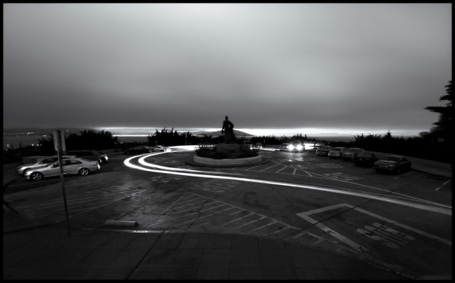 A monochrome picture taken underneath wet, overcast skies from the steps of Coit Tower. 

Car headlights hook around the circular parking lot. A silhouetted Columbus looks out over a gloomy San Francisco Bay in the distance. 