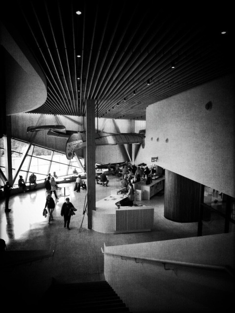 A black and white image of the large entrance lobby of the Canadian Canoe Museum. People can be seen walking and sitting at tables, and large canoes hang from the ceiling into the background.