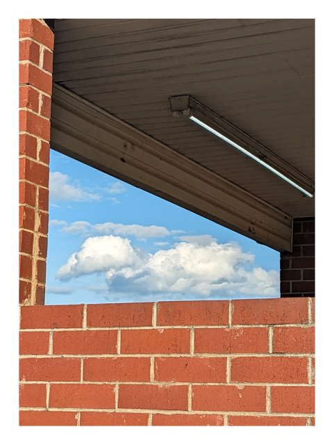 a triangle of a blue sky with big, fancy clouds is visible from outside a red brick car wash stall. a fluorescent bulb on the white plank ceiling inside.