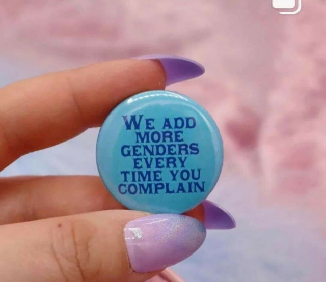 A photo of a light skinned person's hand with manicured nails in lavender. They are holding a blue button that says, "We add more genders every time you complain"