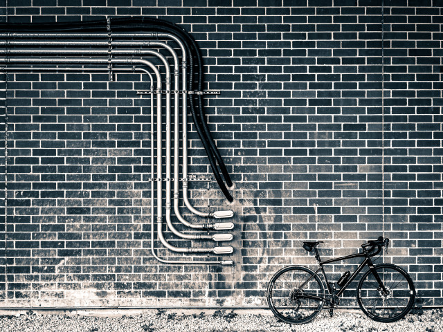 monochrome photo of a black specialized diverge gravel bike, leaning against a brick wall. On the wall, there are five metal electrical conduits, and two black plastic pipes. the conduits and pipes are neatly arranged, and are coming in from the left-hand side of the photo. they make a turn downwards and then another turn to the right where they terminate into the wall just before the bicycle. 