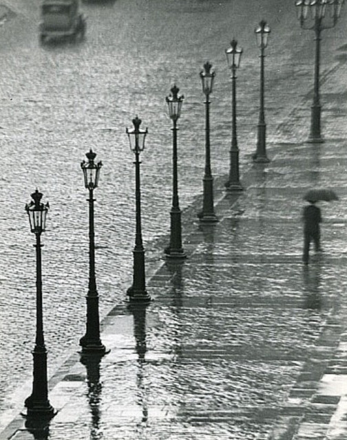 Photography. An old black and white photo of a street and sidewalk covered in rainwater. A man in a black suit and hat is walking with an umbrella across the water-logged square. A row (8) of old street lamps is across the photo. A moving vintage car can be seen in the background. The picture is intentionally somewhat noisy, which reinforces the impression of fleetingness.
Info: Kertesz, Andre (1894-1985) 
was a Hungarian-born photographer known for his groundbreaking contributions to photographic composition and the photo essay. In the early years of his career, his then-unorthodox camera angles and style prevented his work from gaining wider recognition