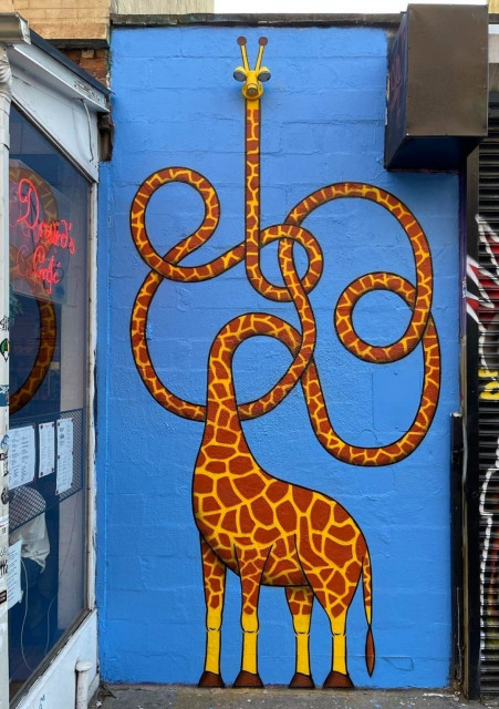 Streetartwall. A giraffe with a very, very long neck was sprayed/painted on a narrow, blue-painted exterior wall. The highlight is what was there before. At the very top was an inconspicuous fixture and two small spotlights at the top pointing in different directions. These spotlights are now the eyes of a giraffe, its neck snaking across the entire wall. I think it's looking at us. Simply marvelous.