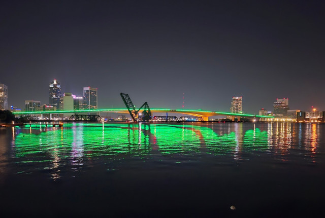 Looking out over a vast river in the darkness of night, the horizon is illuminated with a colorful city skyline and a large concrete bridge spanning the river with a green strip of neon lighting that runs down the center of the bridge, along with the building lights, adds colorful reflections on the river's water below.