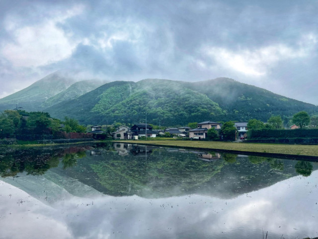 Mountain landscape with mist, greenery, and houses reflected in a tranquil body of water.
