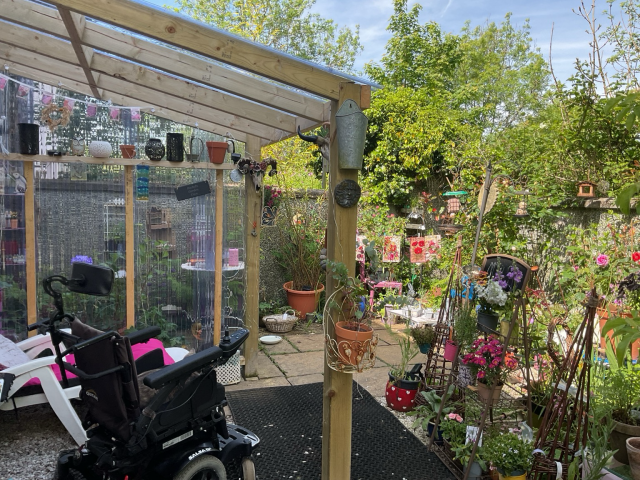 A wheelchair under a shelter with an array of plated pots and shrubs.