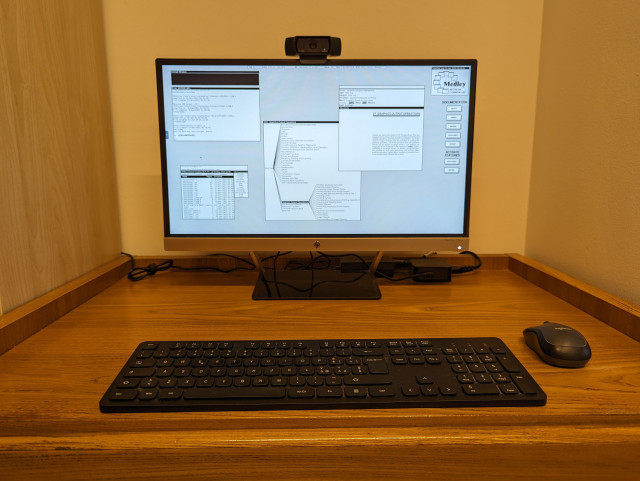A wooden desk with a black keyboard, a black mouse, and a flat LCD monitor with a black webcam on top, The screen shows the black and white desktop of a 1980s graphical workstation environment. The desktop has a gray background pattern and some windows with a white background and a title bar with white text on a black background.