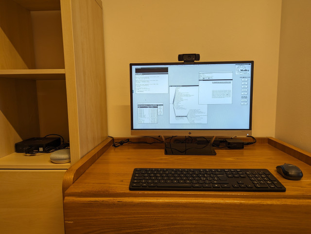 A wooden desk with a black keyboard, a black mouse, and a flat LCD monitor with a black webcam on top, The screen shows the black and white desktop of a 1980s graphical workstation environment. The desktop has a gray background pattern and some windows with a white background and a title bar with white text on a black background. At the left of the desk can be partly seen a wooden bookcase with two shelves. On the bottom shelf are a black computer with a mini PC form factor and a white and gray smart speaker.