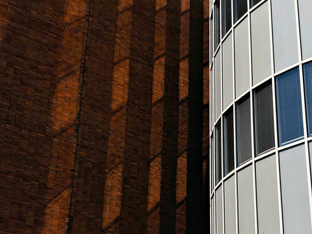 A little of half of the image starting from the left side is a tall brickwall. On the right is a curved section of a buildings with rows of windows and grey panes. Light from the windows is reflected onto the brick wall.
