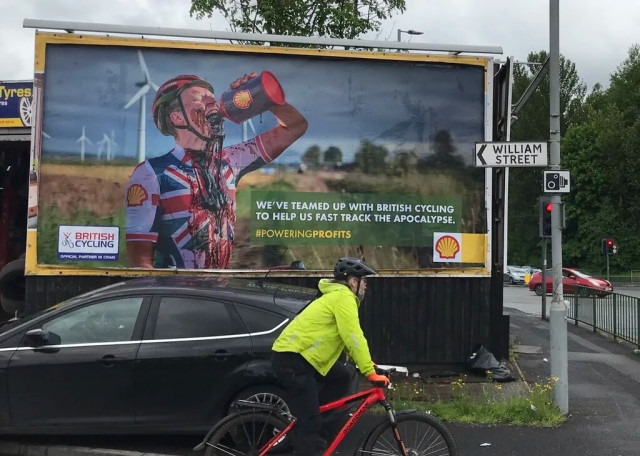 A roadside advertising billboard. Pictured is a cyclist in a union jack jersey drinking a can of Shell oil and spilling it on his chest.
Between the British Cycling and Shell logos it reads ‘We’ve teamed up with British Cycling to help us fast track the apocalypse #PoweringProfits’