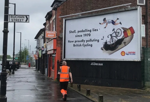 A roadside advertising billboard. Pictured is a cyclist crashing on an oil spill.
In addition to the British Cycling and Shell logos it reads ‘Shell, pedalling lies since 1970. Now proudly polluting British Cycling’.