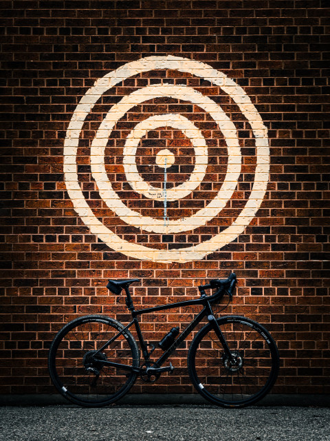 Photo of a black specialized diverge gravel bike standing next to a red brick wall. above the bike is a large bull’s-eye design painted with yellow stripes. 