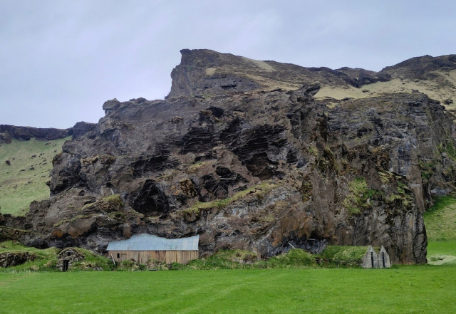 A big volcanic rock surround by green grass. A little barn-like building looks to be halfway swallowed by the rock. The wall facing us is wood in natural tones and the slanted roof is corrugated iron. To the left of it is a tradition stone building where the roof is slanted on both sides, reaching the ground and covered in geeen grass. Far to the right of the frame are to similar stone buildings facing the right.