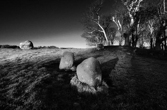 Black and white photo of two small standing stones, aligned to a glacial erratic boulder, close to a clump of bare trees.