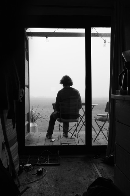 A photo from inside a cabin looking out a patio window/door, where someone is sat at a table working on a laptop, and behind them the world is lost in fog.